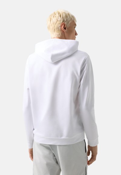 Person with short blond hair wearing a plain white hooded sweatshirt and light gray pants, standing and facing away against a white background.