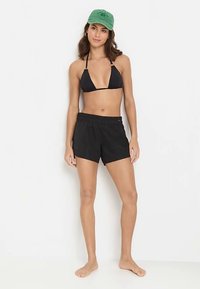 Czarny top bikini z trójkątnymi miseczkami, zestawiony z czarnymi szortami do pływania. Modelka w zielonej czapce, boso na jednolitym tle.