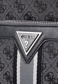 Portofel din țesătură neagră cu un model de logo embosat, accesorii din metal argintiu și un insigne metalic triangular cu "GUESS USA" și un semn de întrebare.