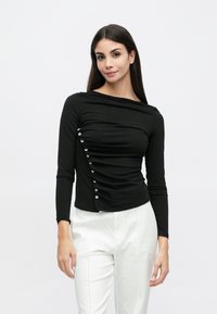 Μαύρο μακρυμάνικο τοπ με λαιμόκοψη σκαφάκι, ρευστή υφή με πιέτες και λεπτομέρειες με κουμπιά στο πλάι, συνδυασ�μένο με λευκό παντελόνι.