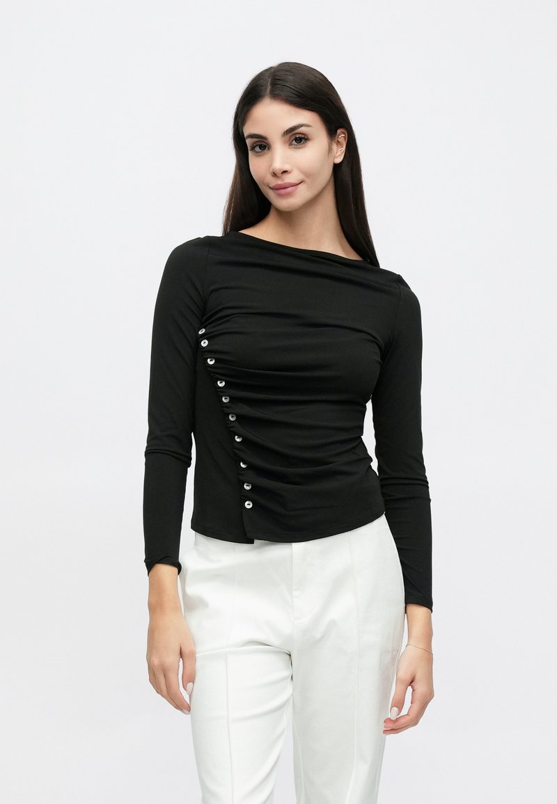 Μαύρο μακρυμάνικο τοπ με λαιμόκοψη σκαφάκι, ρευστή υφή με πιέτες και λεπτομέρειες με κουμπιά στο πλάι, συνδυασμένο με λευκό παντελόνι.