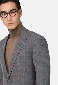 Blazer de cuadros gris con solapas de muesca y cierre de un solo botón. Usado sobre un suéter de cuello alto ajustado marrón. Cuenta con un bolsillo en el pecho.