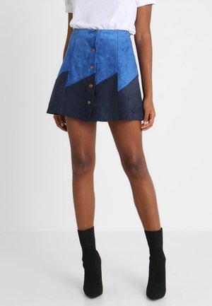 Vrouw draagt een blauwe en marineblauwe mini-rok met knopen aan de voorkant, een wit topje met korte mouwen en zwarte enkellaarsjes, staand tegen een effen achtergrond.