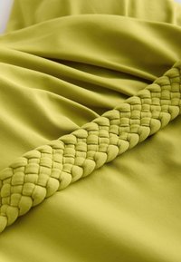 Groene stof met een gladde textuur en een geplooid ontwerp, voorzien van een gevlochten accent in een bijpassende tint langs één rand.