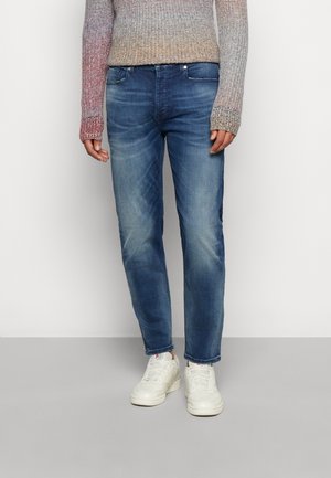 Osoba ubrana w wyblakłe niebieskie dżinsy, białe trampki i wielokolorowy dziergany sweter, stojąca na jednolitym białym tle.