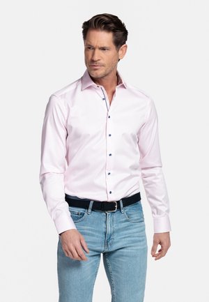 Man draagt een lichtroze overhemd met donkere knopen, ingestopt in lichtblauwe spijkerbroek met een zwarte riem, kijkt naar de zijkant.