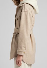 Mujer vestida con abrigo beige con cinturón, con bolsillo en el pecho abotonado y puños, sobre sudadera y pantalones blancos, vista de lado.