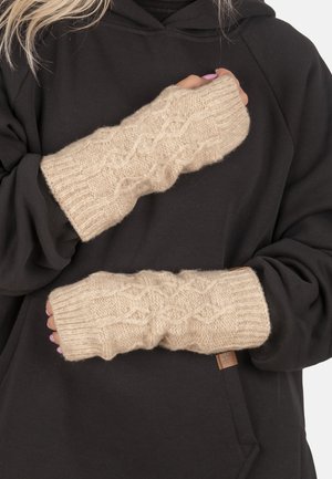 Guanti senza dita beige lavorati a maglia con motivo a treccia indossati sopra un indumento nero a maniche lunghe, mani incrociate sul petto.