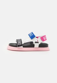 Valkoiset litteät sandaalit mustalla pohjalla, vaaleanpunaisella ulkopohjalla ja säädettävillä mustilla, sinisillä ja vaaleanpunaisilla strap-on-hihnoilla, joissa on valkoista tekstibrändäystä.