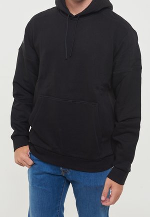 Person iført en ensfarvet sort hættetrøje med frontlomme og snore, kombineret med blå denimjeans.