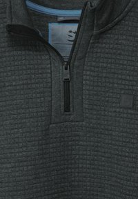 Svart zip-up sweatshirt med ett texturerat mönster, har en hög krage, blå innerfoder och en logotyp på vänster bröst.