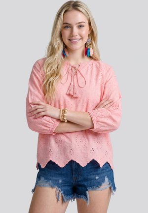 Giovane donna con lunghi capelli biondi indossa una camicetta rosa traforata, pantaloncini di jeans, orecchini colorati con piume e braccialetti di perline, con le braccia incrociate, sorridente.
