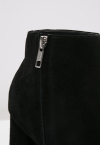 Bottine en daim noir avec une fermeture éclair métallique YKK, une texture lisse et une silhouette ajustée avec des détails de coutures le long du côté.