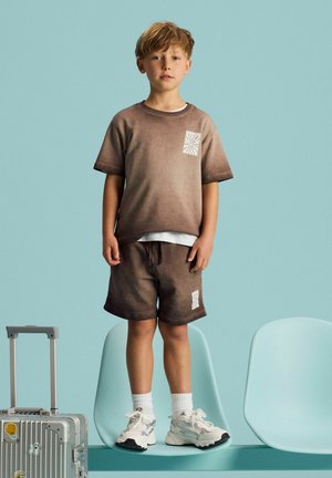 Garçon portant un short marron et un t-shirt avec des baskets blanches, debout sur une chaise bleue à côté d'une valise argentée avec des autocollants sur fond bleu clair.