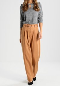 Szara bluzka z długim rękawem, z dekoltem w kształcie łezki i bufkami na ramionach, zestawiona z wysokim stanem, plisowanymi spodniami w kolorze karmelowym oraz czarnymi szpilkami.