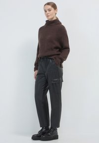 Maglione marrone lavorato a maglia con colletto alto, abbinato a pantaloni cargo neri lucidi con tasche laterali e cintura elastica. Stivali neri con lacci.