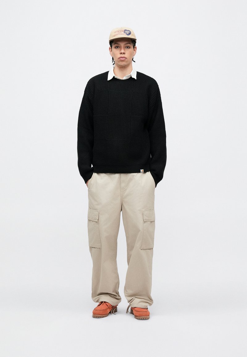 Sort strikket sweater, beige cargobukser med sidelommer, orange sko og en beige kasket med logo. Simpelt, afslappet tøj.