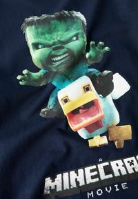Minecraft-inspirert T-skjorte med en grønn karakter med sint uttrykk og en tegneserieand, trykt på marineblått stoff, med hvit tekst under.