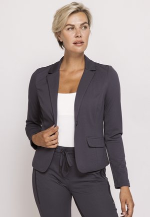 Blonde Frau trägt ein dunkelgraues Sakko und passende Hose über einem weißen Oberteil, schaut mit neutralem Ausdruck zur Seite.