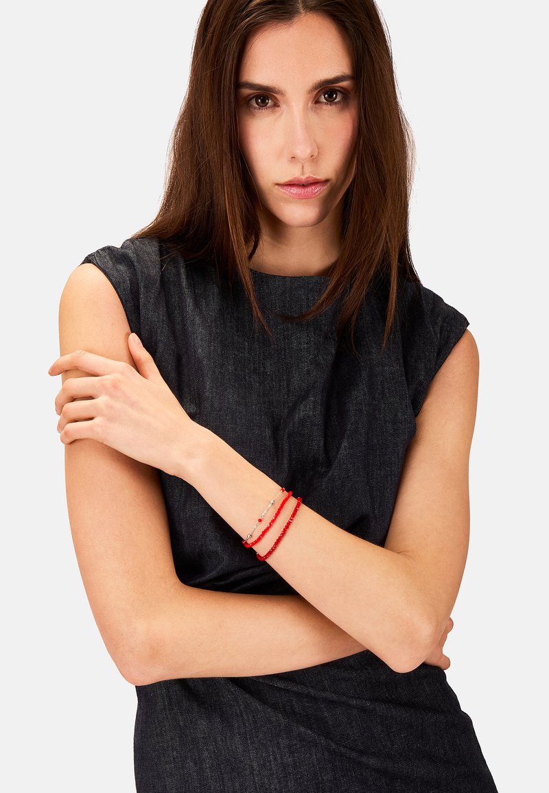 Bracciale di perline rosse con accenti d'argento, composto da tre fili di perline tonde. Indossato su un polso, completa un top senza maniche di colore scuro.