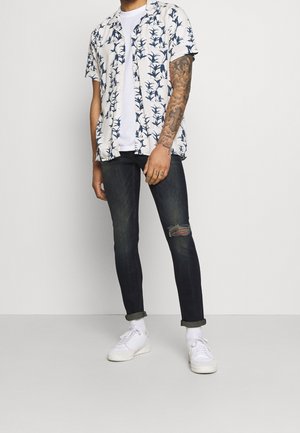 Camicia bianca a maniche corte con motivo floreale blu, indossata sopra una maglietta bianca semplice. Abbinata a jeans scuri e sneakers bianche.