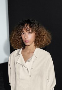 Lichtbeige oversized shirt met een kraag, voorzien van een gedeeltelijke knoopsluiting en een decoratieve veiligheidsspeldaccent. Het model heeft krullend haar.