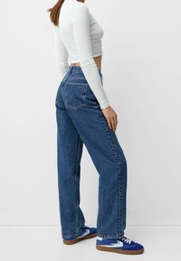 Höga blå denimjeans med rak benvidd, med en klassisk fem-fickorsdesign och fransad nederkant. Bärs med en vit långärmad topp.
