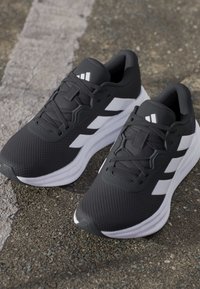 Paire de chaussures de course athlétiques noires et blanches avec lacets sur une surface asphaltée texturée.
