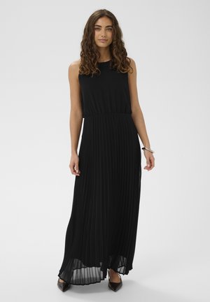 Vrouw met krullend haar, gekleed in een zwarte mouwloze geplooide maxi-jurk en zwarte puntige schoenen, staand op een effen lichtgrijze achtergrond.