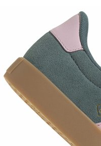 Grön mocka sneakersula med rosa läderaccent och texturerad brun gummisul, som visar sömnadsdetaljer och tygtextur.