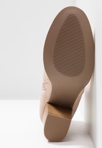 Chaussure à talon haut en cuir suédé rose clair avec un talon en bois. La semelle présente des rainures horizontales pour une meilleure adhérence. Couture visible sur les côtés.