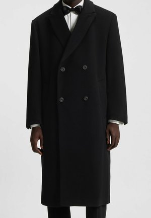 Manteau noir à double boutonnage porté sur une chemise blanche avec un nœud papillon noir, présenté sur une personne debout, du cou aux genoux.