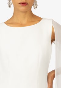 Witte jurk met een bateauhals, doorschijnende lange mouwen en een gladde stof. Model draagt ornamentale zilveren oorbellen.