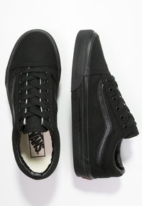 Paire de baskets Vans noires avec lacets noirs, logo visible à l'intérieur et bande latérale distinctive, vues de dessus et de côté.