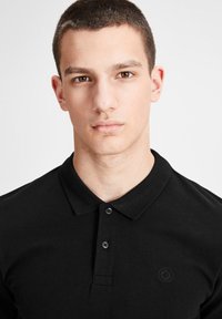 Polo noir en tissu texturé, avec une patte de boutonnage à trois boutons et un logo discret sur la poitrine gauche. Design simple et classique.