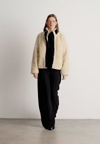 Μπεζ ψεύτικο γούνινο μπουφάν με κομμένη σχεδίαση, ανοιχτό μπροστά, υφή στην επιφάνειά του, φορεμένο πάνω από ένα μαύρο σύνολο. Το μοντέλο ποζάρει σε μια μινιμαλιστική ρύθμιση.