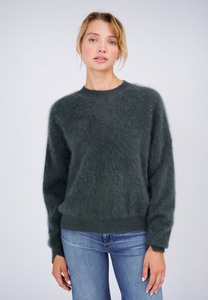 Frau, die einen dunkelgrünen, flauschigen Pullover und blaue Jeans trägt, steht vor einem einfarbigen hellen Hintergrund.