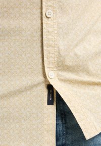 Camisa de manga corta de tela amarillo claro con un patrón geométrico, que presenta un botón blanco y una etiqueta de marca visible sobre un denim oscuro.
