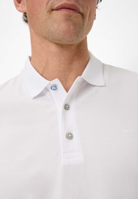 Polo bianco realizzato in un tessuto testurizzato, con collo a righe, due bottoni grigi e blu, e un logo discreto sul petto.