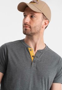 Šedé triko s krátkým rukávem typu henley se žlutým knoflíkovým zapínáním. Kšiltovka v béžovém odstínu s logem vyšitým na přední straně.