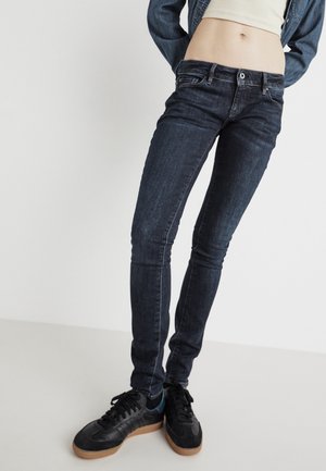 Personne portant un jean skinny foncé, des baskets noires avec des semelles en gomme, un top court blanc et une veste en jean bleue sur un fond uni.