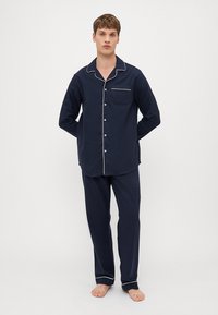 Pijamas em algodão azul marinho com uma camisa de botão com um bolso de remendo e com detaques brancos, acompanhados por calças combinando. Calçado não incluído.