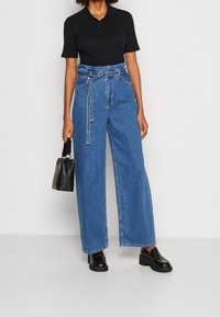 Kvinna i en svart kortärmad topp, högmidjade vida blåjeans med bälte, svarta loafers och håller en liten svart handväska.