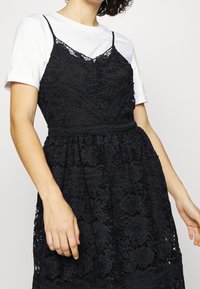 Robe noire en dentelle avec motif floral, bretelles spaghetti ajustables et taille cintrée. Le design en couches donne une apparence texturée.