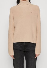 Beżowy sweter z ribbowanym kołnierzem i długimi rękawami, noszony z czarnymi spodniami o wysokim stanie, z małym czerwono-białym-niebieskim logo na klatce piersiowej.