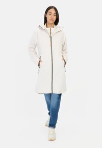 Leichter, langer beiger Mantel mit Kapuze, Vorderreißverschluss und Holzakzenten. Verfügt über Seitentaschen und ist mit blauen Jeans kombiniert.