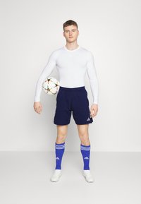 Witte lange mouwen sporttop gecombineerd met marineblauwe shorts en blauwe sokken met witte strepen, terwijl hij een voetbal vasthoudt, met Adidas-logo.