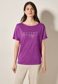 T-shirt court violet à manches courtes avec une coupe ample, comportant un imprimé graphique en texte blanc et gris. Tissu doux et léger avec une apparence texturée.