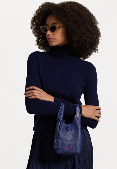 Jūras spēku turtlenecks džemperis un plīvurēta svārki, turot nelielu zilu teksturētu ādas somu ar šuvju akcentiem un logotipa detaļu.