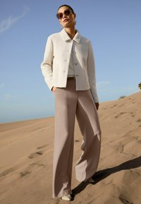Frau trägt Sonnenbrille, weiße strukturierte Jacke und beige weit geschnittene Hose, steht auf sandigen Dünen unter einem klaren blauen Himmel.